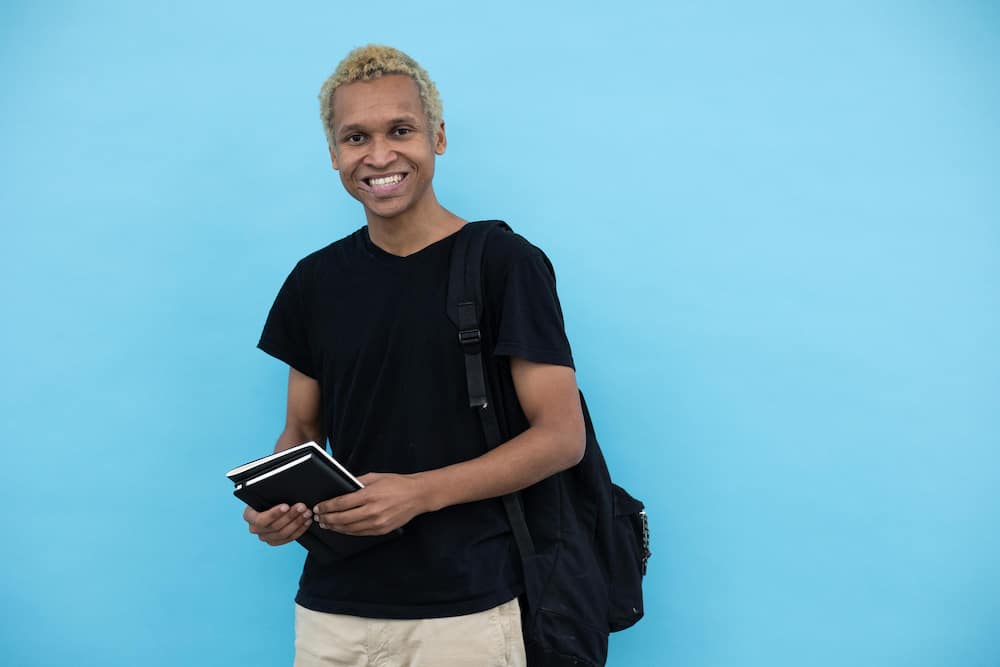 A young man in a black tee is holding books and carrying a backpack