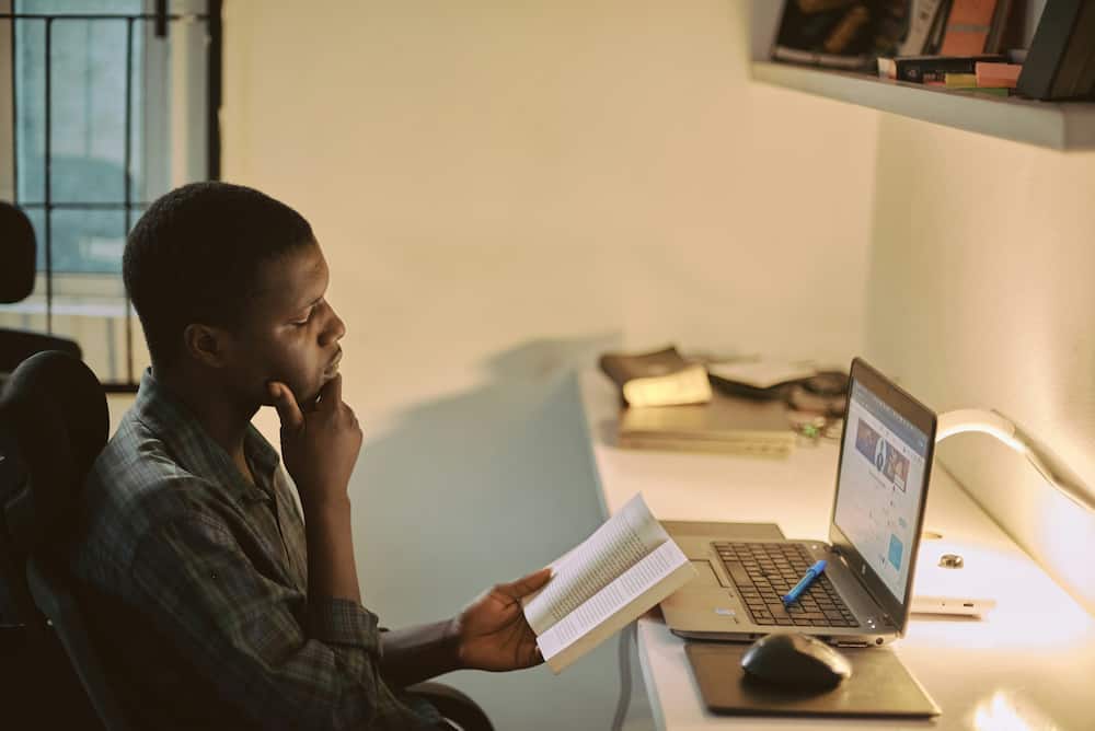 A young man in a checkered shirt is reading a book in front of a laptop